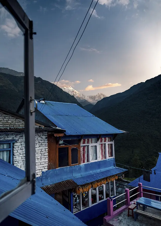 Blue-roofed mountain lodge framed by an open window beneath the Himalaya in Nepal